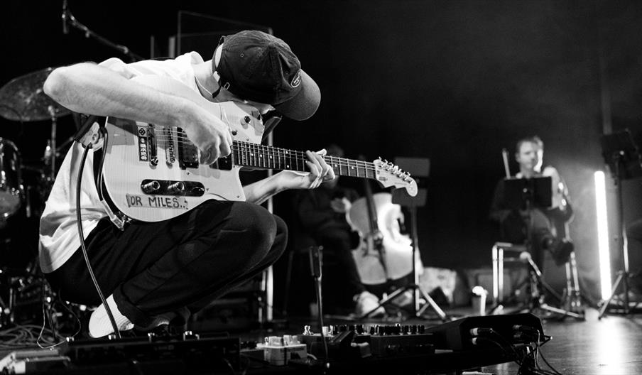 Black-and-white image of guitarist Rory Friers crouching low on stage over a pedalboard while playing an electric guitar covered in stickers.