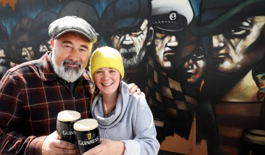 A man and women stood infront of a large mural of people, each holding a pint of Guiness. The man is wearing a check shirt and a grey flat cap and the