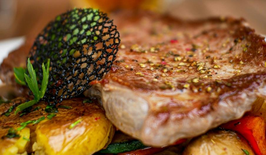 Close-up of a grilled steak topped with herbs and seasoning, served on a bed of roasted vegetables and potatoes, with a decorative crisp garnish on to