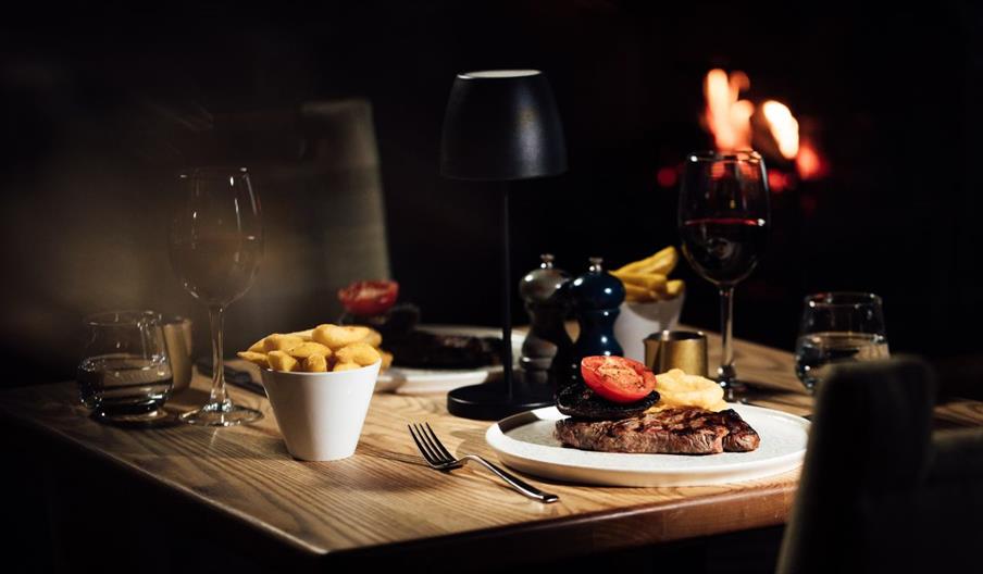 This image shows a low lit restaurant with steak dishes sitting on the table accompanied by chips and glasses to wine beside a fire