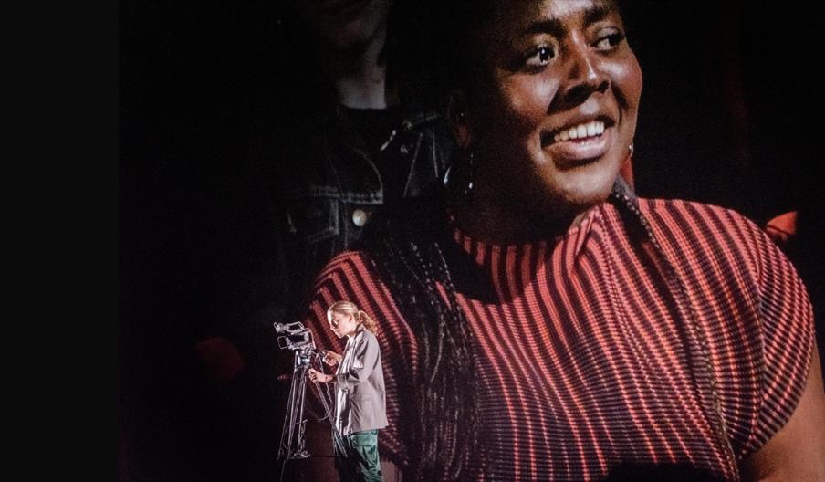 A smiling Black woman wearing a red-and-black striped top, with a smaller image in the bottom left of a person operating a professional camera mounted