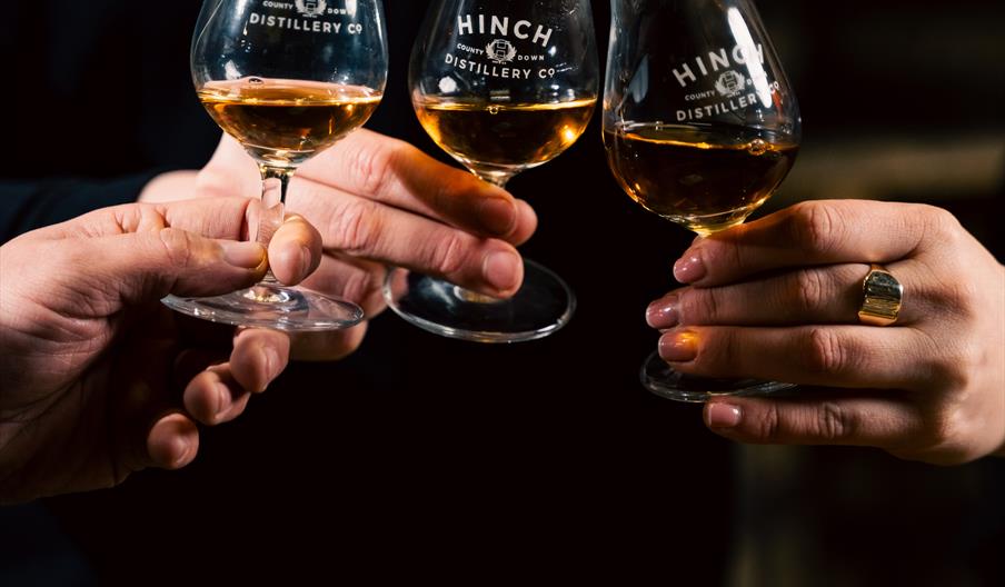 Three people holding small tasting glasses of amber whiskey labelled "Hinch Distillery Co." over a wooden barrel.