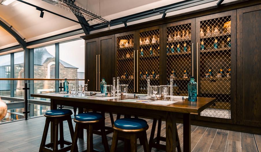 whiskey labelled "Hinch Distillery Co." over a wooden barrel.  A modern tasting room or lab space with a long wooden table set for a spirits or-tastin