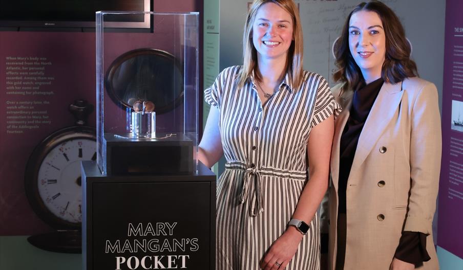 Two women standing beside a display case containing Mary Mangan's pocket watch at Titanic Belfast, with exhibition panels about Mary and the Addergool
