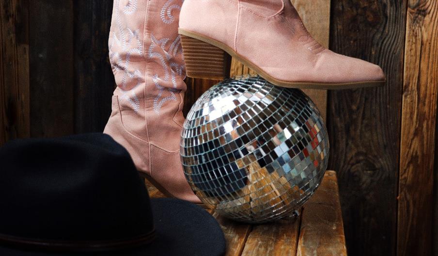 A pink cowboy boot resting on a disco ball with a cowboy hat beside it