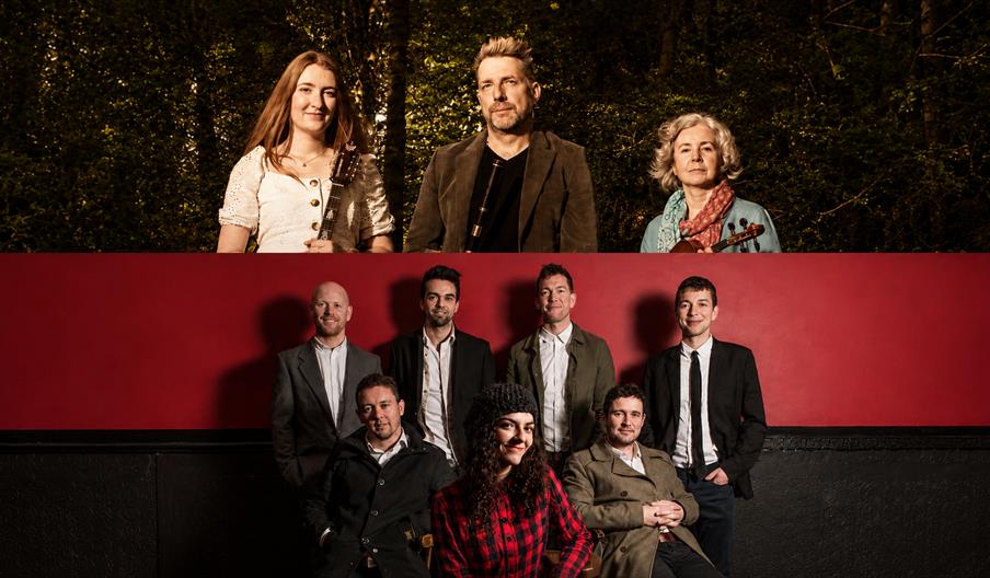 A split promotional image featuring two groups of musicians: the top half shows three performers standing outdoors in warm sunlight against a wooded b