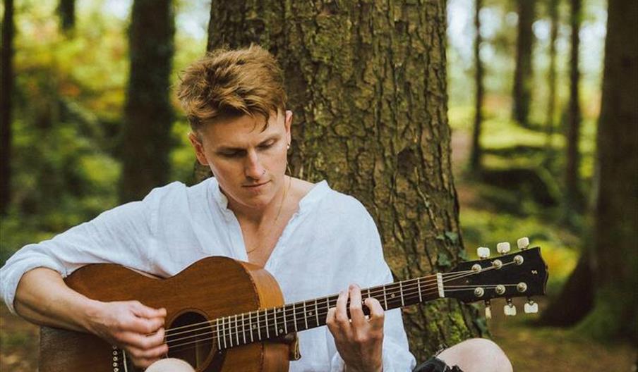 Seán Ó Meara sits next to a tree with his guitar