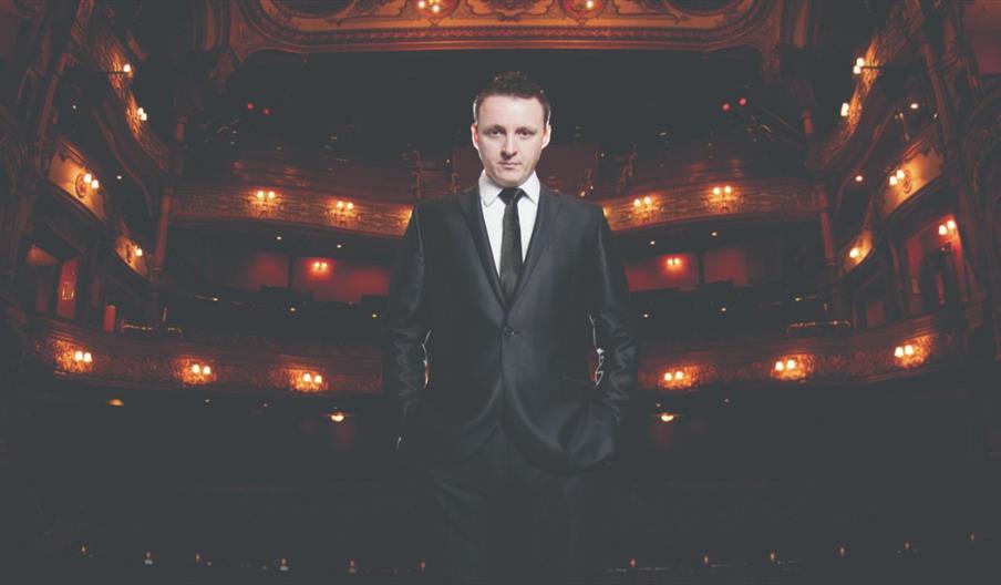 A man in a suit and tie stands on stage with his back to an empty auditorium looking at the camera