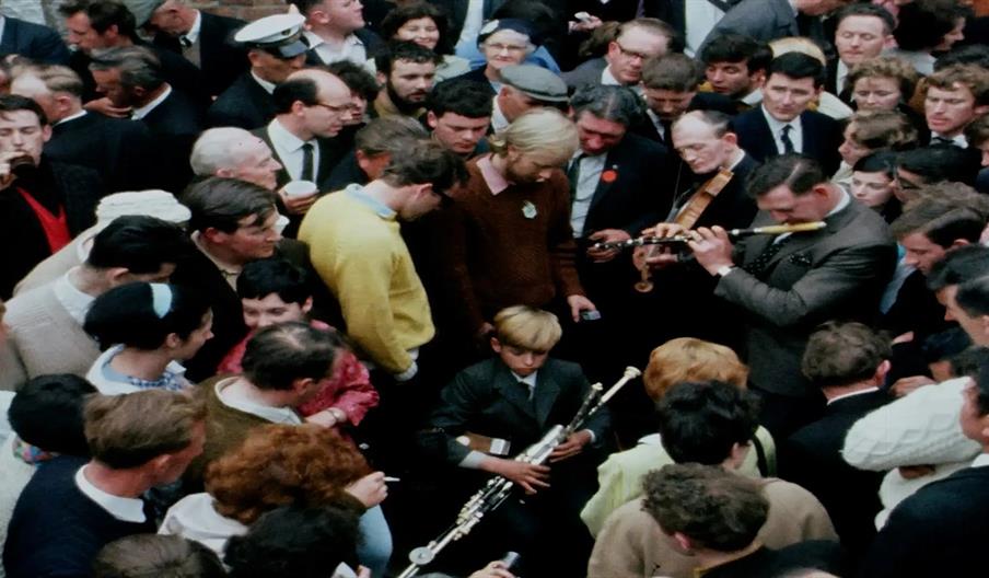 1960s colour photograph of a street scene from a Fleadh Cheoil na hÉireann event. The street is packed full of people and a young boy with an uillean