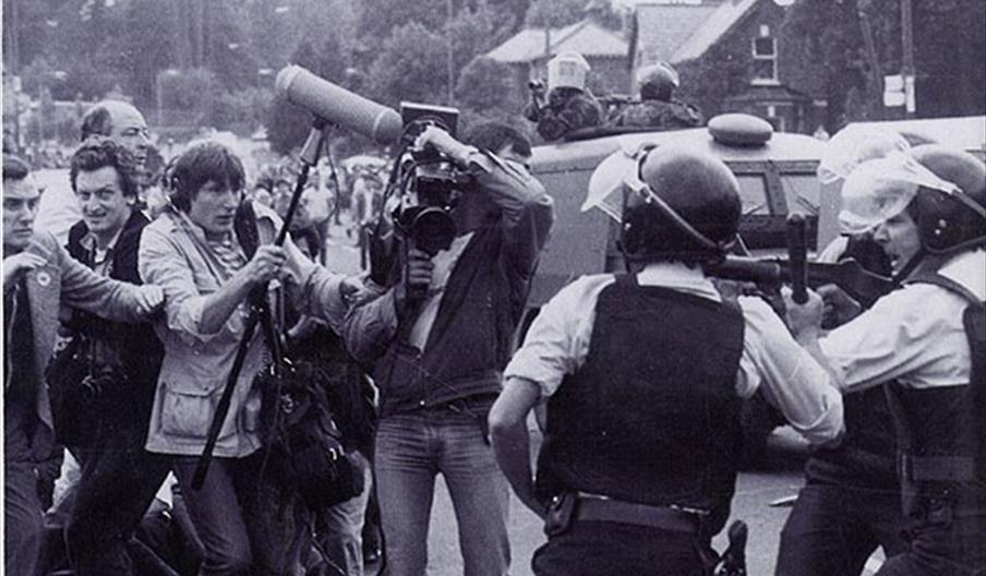 A group of camera journalists look on with concern as police officers wearing helmets and firearms vests point baton firearms past them