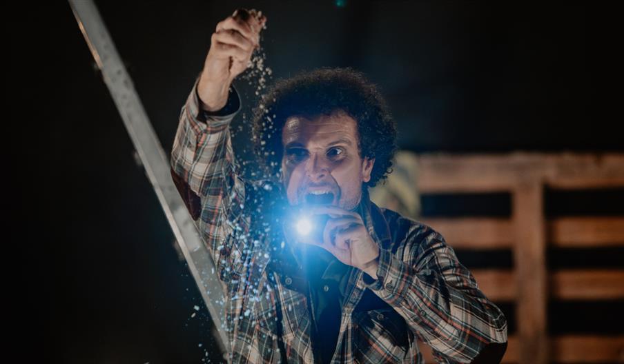 A man with dark curly hair in a plaid shirt holds a bright flashlight near his mouth with one hand while sprinkling sand or grain overhead with the ot