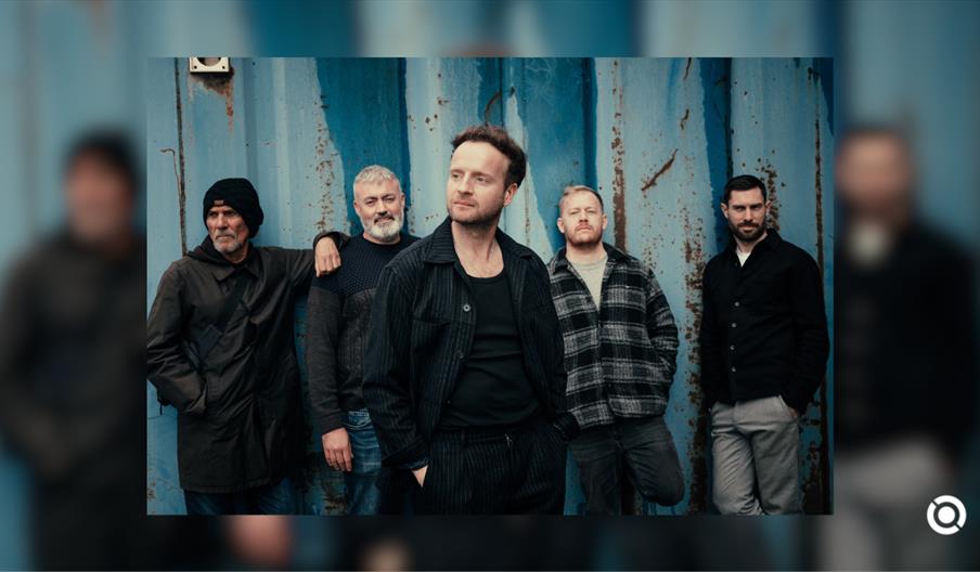 A group promotional photograph featuring five men standing side-by-side outdoors against a weathered, vertically corrugated blue metal wall.  The indi