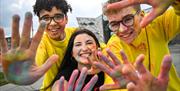 A close up shot of three young people smiling at the camera, their hands messy with colour powder.