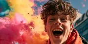 A young man laughs at the camera while a plume of colour powder rises behind him.