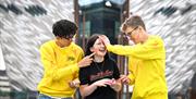 Three young people throw color powder at each other, with Titanic Belfast in the background.