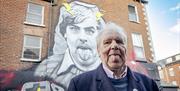 Picture shows Terri Hooley standing in front of a mural depicting himself with his tongue out