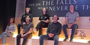 Group of people wearing headsets seated on tiered wooden benches inside an exhibition space with the words 'Life on The Falls would never be the same'