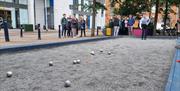 Picture shows petanque piste which is a blue rectangle with gravel and some silver ptanque boules on it. Somebody in the distance throws a boule