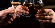 Three people holding small tasting glasses of amber whiskey labelled "Hinch Distillery Co." over a wooden barrel.