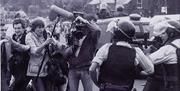 A group of camera journalists look on with concern as police officers wearing helmets and firearms vests point baton firearms past them
